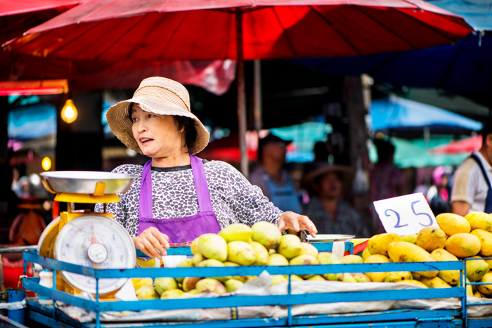 Documentary photography of a woman standing behind a basket of fruit at a busy market in Chiang Mai, Thailand