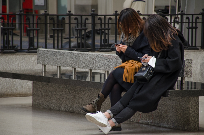 Street photo of two girls sitting on a bench and using smartphones, demonstrating mid and dark tones in images